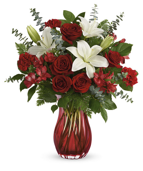 A glass vase holds a bouquet of red roses, white lilies, red alstroemeria, and greenery, standing against a white background.