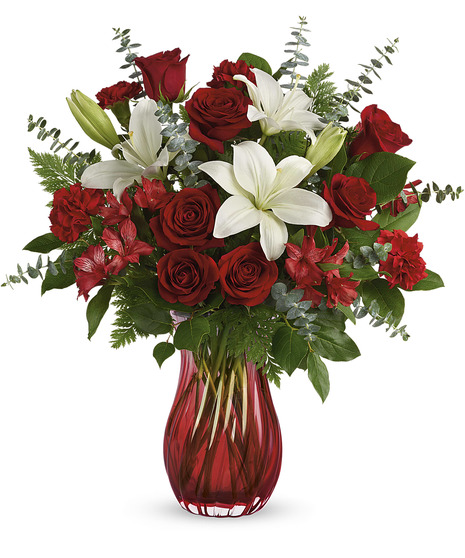 A glass vase holds a bouquet of red roses, white lilies, red alstroemeria, and greenery, standing against a white background.