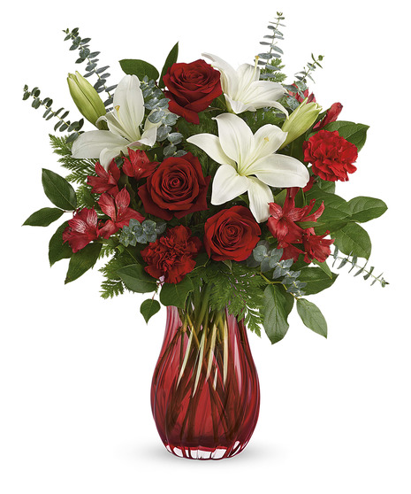 A glass vase holds a bouquet of red roses, white lilies, red alstroemeria, and greenery, standing against a white background.