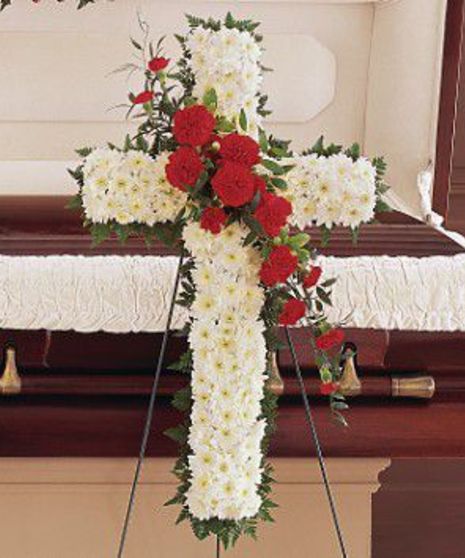A cross-shaped floral arrangement of white and red flowers stands in front of a closed wooden casket with a white cloth draped over it