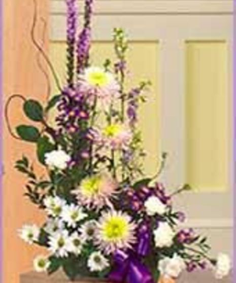 Floral arrangement of assorted white, purple, and yellow flowers, adorned with a purple ribbon, situated indoors against a light-colored paneled wall.