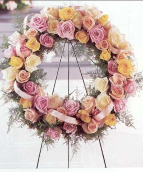 A wreath made of pink, yellow, and white roses adorned with greenery and ribbons sits on a black metal stand in a light-colored indoor setting.