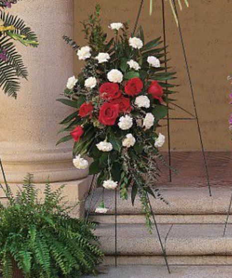 Floral arrangement of red and white flowers on a stand, situated in front of beige columns and steps with green ferns nearby.