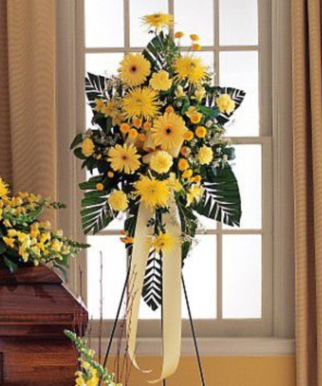 A large, yellow flower arrangement stands on a black tripod in front of a tall window with sheer white curtains
