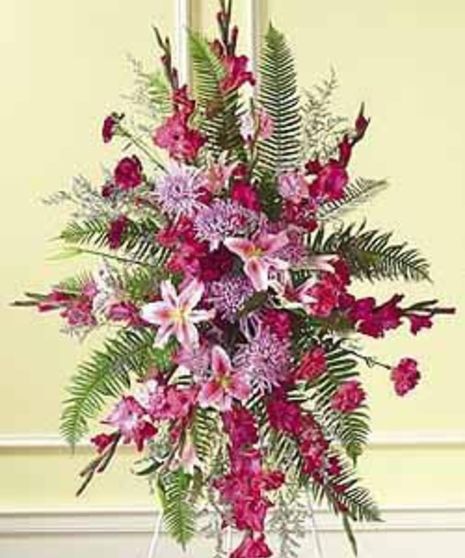 A vibrant floral arrangement features pink lilies, red carnations, and green ferns, displayed indoors against a pale yellow wall with white trim.
