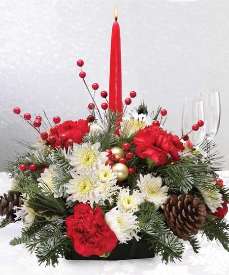 A red candle surrounded by red and white flowers, pine branches, pinecones, red berries, and gold ornaments arrange in a festive centerpiece on a white tablecloth.