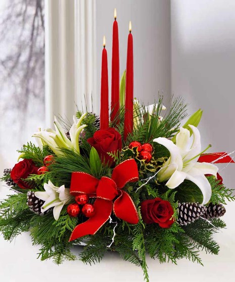 A floral arrangement featuring three lit red candles, red roses, white lilies, pinecones, and green foliage, adorned with a large red bow, placed indoors near a window.
