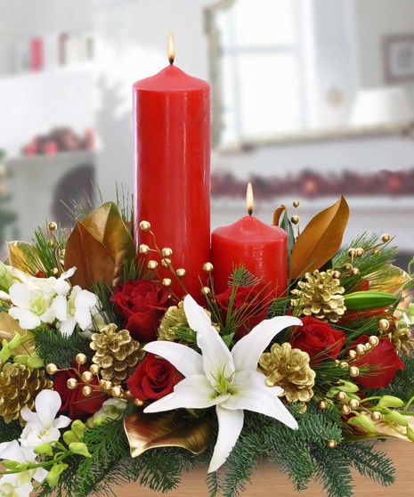 Red candles burning among festive decorations featuring white lilies, red roses, golden pine cones, and green foliage. 