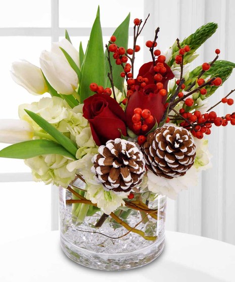 A clear vase holds white tulips, red roses, pinecones, and red berries, arranged with green leaves and branches