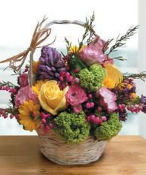 Basket of colorful flowers including roses, daisies, and hydrangeas on a wooden table, placed near a window with light streaming in.