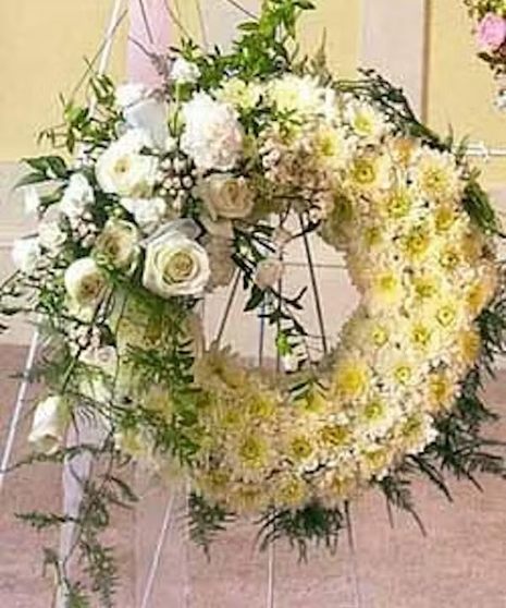 A floral wreath composed of white and yellow flowers, including roses and daisies, adorned with greenery, is displayed on an easel stand