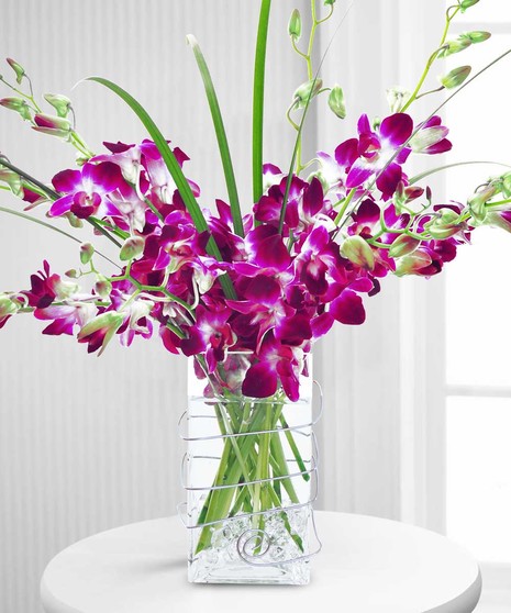 A bouquet of purple and white orchids arranged in a clear, rectangle vase on a small, round table in a bright room with white walls and a window.