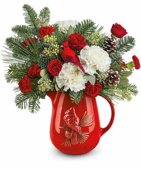 A festive arrangement of red roses, pinecones, and lush greenery sits in a red vase adorned with a cardinal bird design, complemented by white flowers and red berry accents.