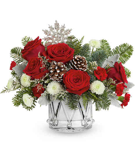 A festive arrangement of red roses, pinecones, and lush greenery sits in a red vase adorned with a cardinal bird design, complemented by white flowers and red berry accents.