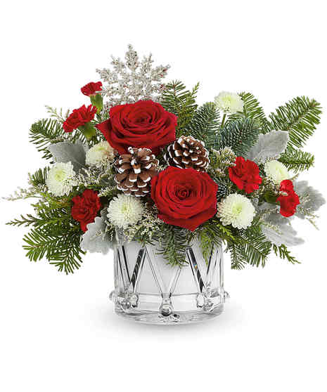 A festive arrangement of red roses, pinecones, and lush greenery sits in a red vase adorned with a cardinal bird design, complemented by white flowers and red berry accents.