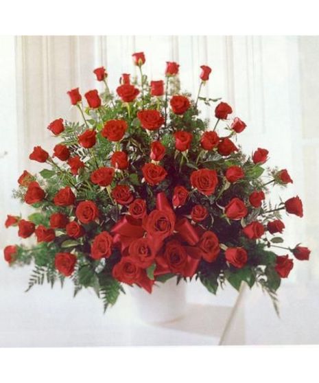 A bouquet of numerous red roses with green foliage arranged in a white vase, adorned with a red bow, set against a light background.