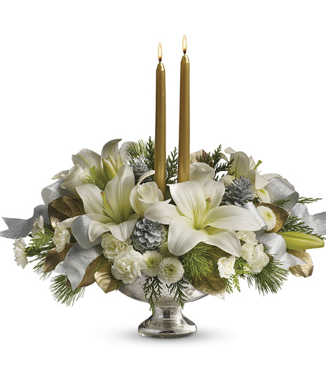 A silver vase holds a holiday bouquet of white lilies, pinecones, and greenery, accentuated by two lit gold candles and decorative silver and gold leaves.