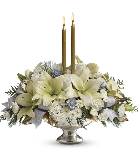 A silver vase holds a holiday bouquet of white lilies, pinecones, and greenery, accentuated by two lit gold candles and decorative silver and gold leaves.