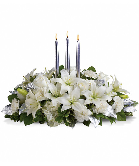 Three lit silver candles arranged within a bouquet of white lilies, carnations, roses, and green foliage with silver leaves, set against a plain white background.