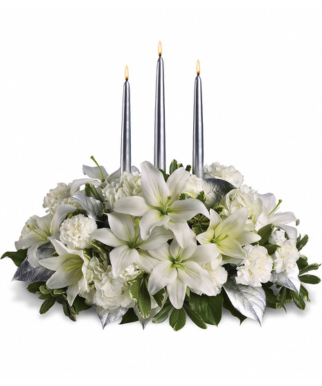 Three lit silver candles arranged within a bouquet of white lilies, carnations, roses, and green foliage with silver leaves, set against a plain white background.