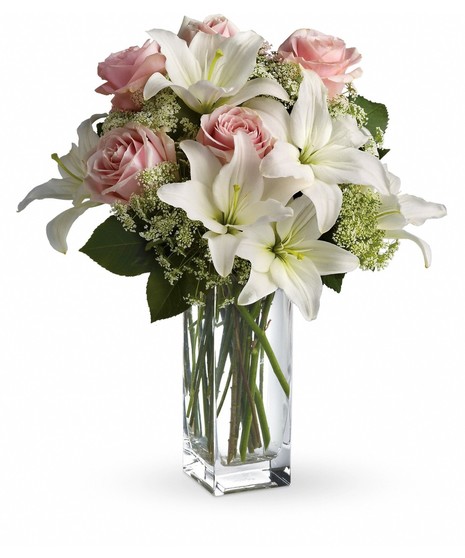 A bouquet of white lilies and pink roses sits in a tall, clear glass vase, surrounded by green leaves and white filler flowers, against a plain white background.