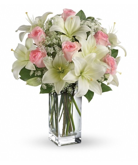 A bouquet of white lilies and pink roses sits in a tall, clear glass vase, surrounded by green leaves and white filler flowers, against a plain white background.