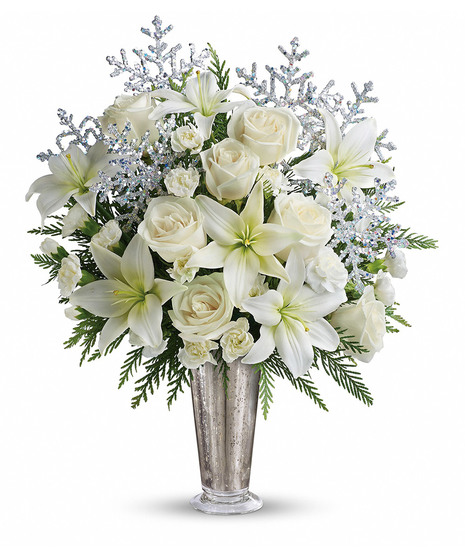 A silver vase holds white roses, carnations, and greenery, adorned with glittery, silver snowflake decorations, set against a plain white background.