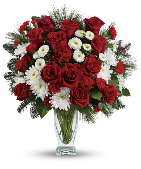 A crystal vase holds a bouquet of vibrant red roses, carnations, white daisies, and pine branches, arranged elegantly against a plain white background.