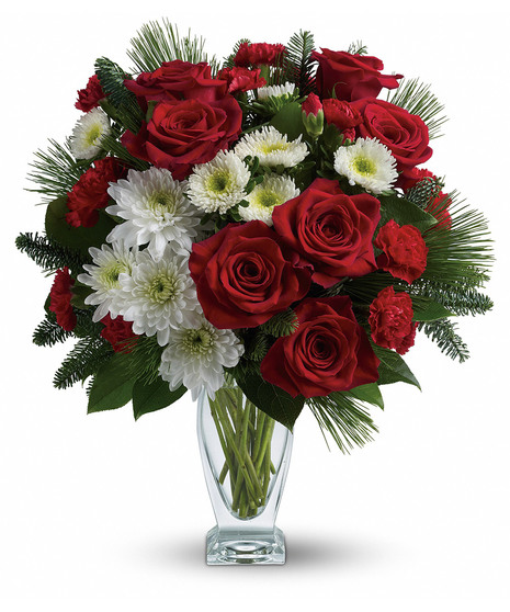 A crystal vase holds a bouquet of vibrant red roses, carnations, white daisies, and pine branches, arranged elegantly against a plain white background.