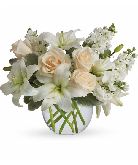 A clear round vase holding a bouquet of white lilies, cream roses, and small white flowers, surrounded by green leaves, against a plain white background.