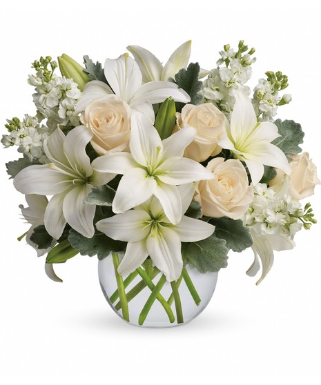 A clear round vase holding a bouquet of white lilies, cream roses, and small white flowers, surrounded by green leaves, against a plain white background.