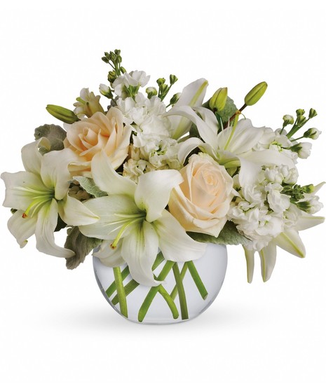 A clear round vase holding a bouquet of white lilies, cream roses, and small white flowers, surrounded by green leaves, against a plain white background.